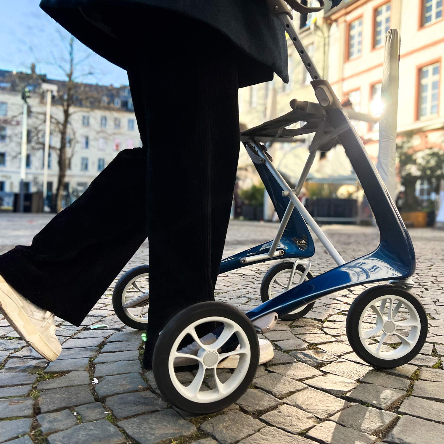 A person in black pants and white shoes walks on a cobblestone street using a byACRE Carbon Ultralight Rollator, with buildings and trees in the background.