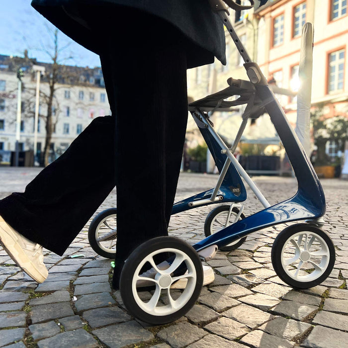 A person in black pants and white shoes walks on a cobblestone street using a byACRE Carbon Ultralight Rollator, with buildings and trees in the background.