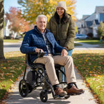 An older man smiles while seated in a Journey Air Elite Max Ultra-Lightweight Portable Power Chair on a sunny sidewalk, as a younger woman in a coat and beanie stands behind him. Autumn leaves and suburban homes can be seen in the background.
