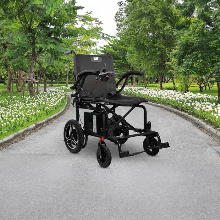The Journey Air 2026 Portable Folding Electric Power Chair, with a 300 lb weight capacity, is placed on a paved path in a park bordered by green trees and white flowers.