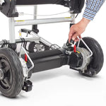 Hand adjusting a red lever near the rear wheel of the Journey Zoomer lightweight folding power chair, demonstrating the brake release or locking mechanism with visible frame and wheel detail.