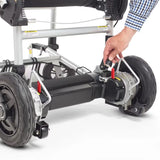 Hand adjusting a red lever near the rear wheel of the Journey Zoomer lightweight folding power chair, demonstrating the brake release or locking mechanism with visible frame and wheel detail.