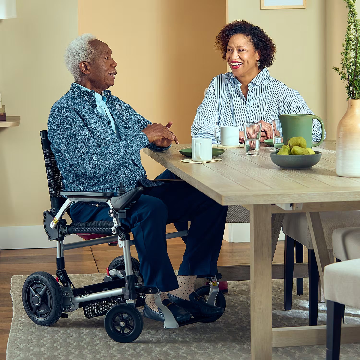 Older man using a Journey Zoomer lightweight folding power chair while sitting at a wooden dining table with a woman, both smiling and chatting over coffee in a bright, cozy indoor setting.