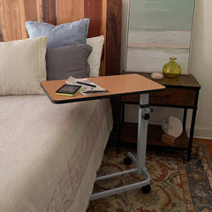 A mobile over-bed lap desk with a light wood top and silver metal frame on casters, shown resting on a bed in a bedroom next to a nightstand.