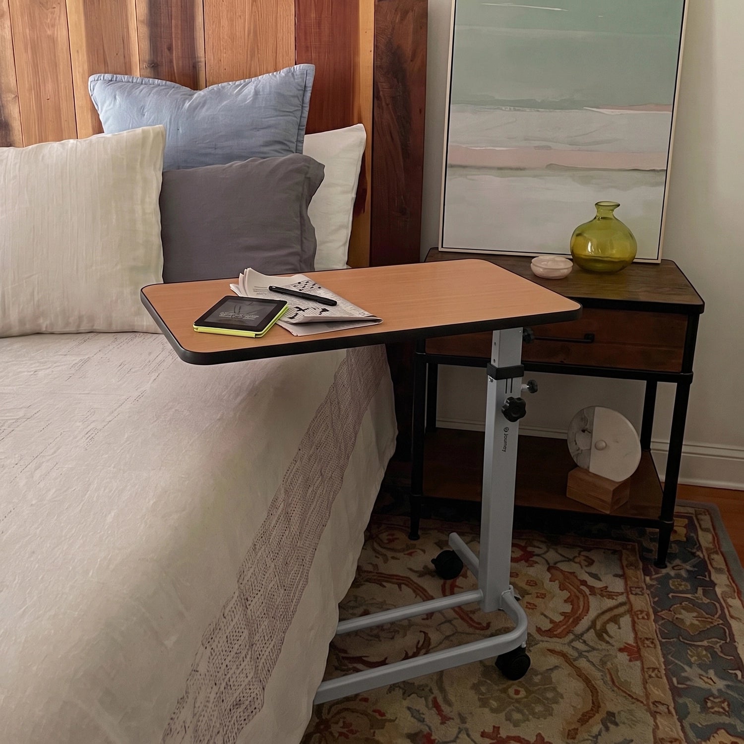 A mobile over-bed lap desk with a light wood top and silver metal frame on casters, shown resting on a bed in a bedroom next to a nightstand.