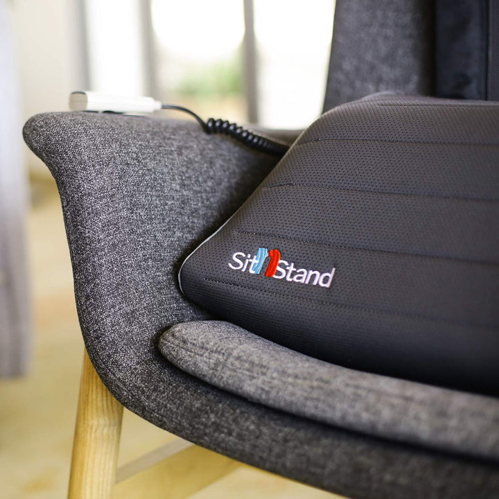 A close-up of a grey fabric chair with a black cushioned seat labeled SitNStand Lift Assist Classic for Home, featuring a portable lift assist cushion and a wired remote control resting on the armrest.