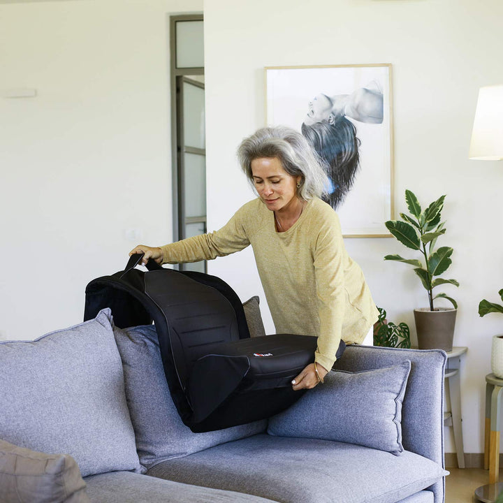 A woman with gray hair places the SitNStand Lift Assist Classic for Home onto a gray sofa in a bright living room decorated with plants and framed artwork.