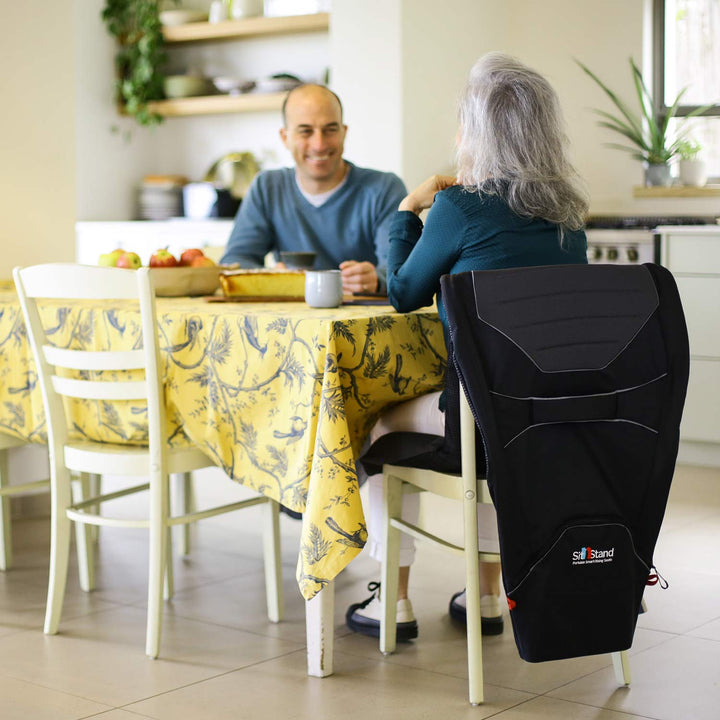 A man and an older woman with gray hair sit at a kitchen table. The woman uses a black back support and the SitNStand Lift Assist Classic for Home cushion on her chair while they have a conversation.