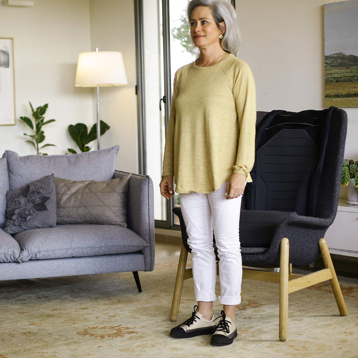 A woman with gray hair stands indoors next to a black SitNStand Lift Assist Classic for Home, wearing a yellow long-sleeve top, white pants, and black-and-white sneakers. Behind her are a gray sofa, lamp, and plants.