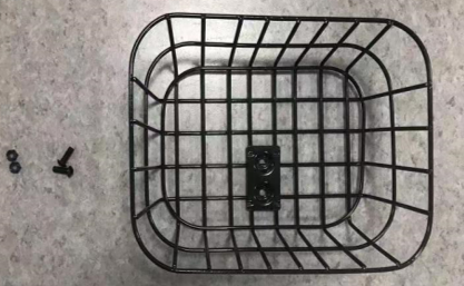 The So Lite® Scooter Basket Sub Assembly, a black metal wire basket with a center mounting bracket, rests on a gray countertop. Two small screws and two nuts are placed to the left of the basket.