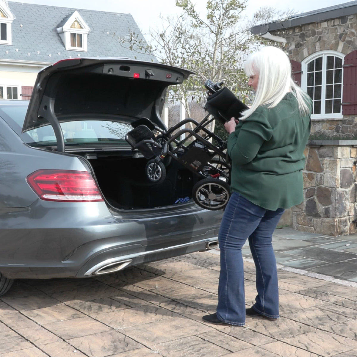 A lady with gray hair loading a Journey Power Chair into the trunk of a car outside a house.
