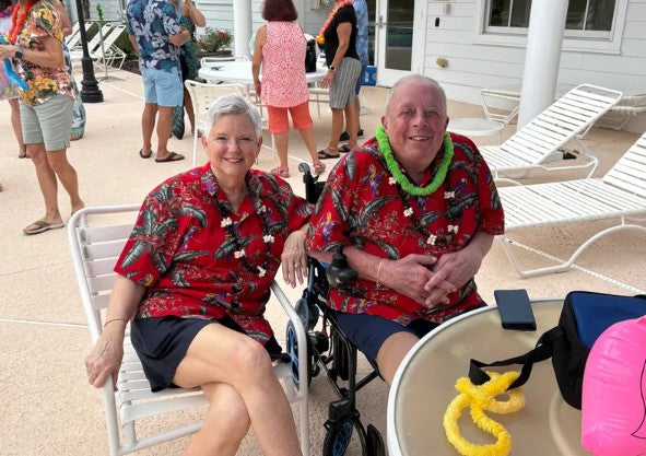 Two people in matching red shirts with tropical patterns sitting one sitting  a lounge chair and one sitting in a Journey Air Elite power chair by a poolside.
