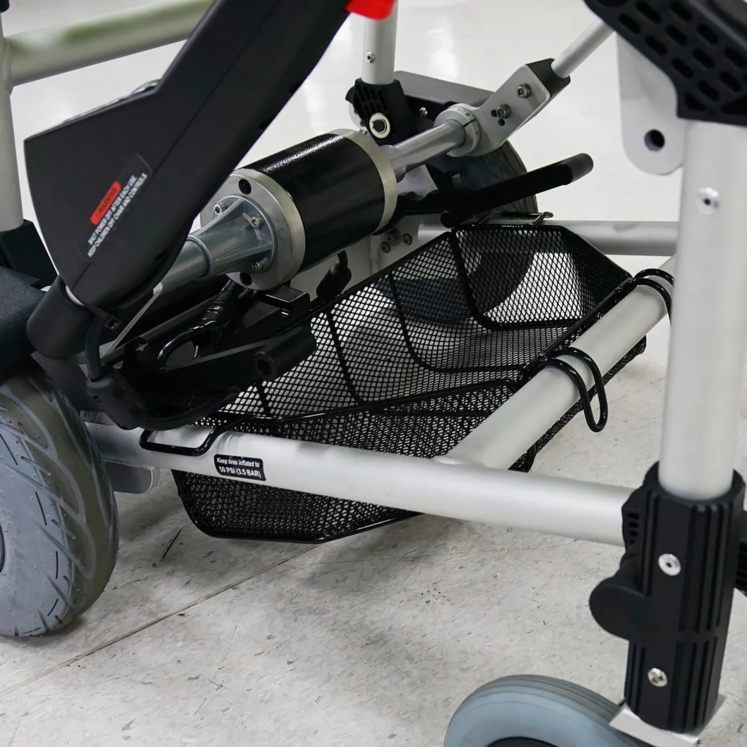 Close-up of the Journey Zinger Power Chair Basket – Portable & Strong, attached under the Zinger Power Chair’s sturdy metal frame and between its wheels, ideal for carrying mobility scooter accessories. The chair sits on a white tiled floor.