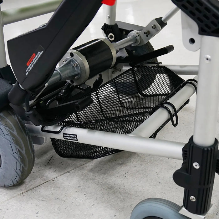 Close-up of the Journey Zinger Power Chair Basket – Portable & Strong, attached under the Zinger Power Chair’s sturdy metal frame and between its wheels, ideal for carrying mobility scooter accessories. The chair sits on a white tiled floor.