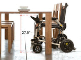 The Journey Zoomer Lightweight Folding Power Chair fits under a wooden dining table with a 27.5-inch height, as shown by a red arrow. Plates are stacked on the tabletop, highlighting the chair’s compatibility for dining use.