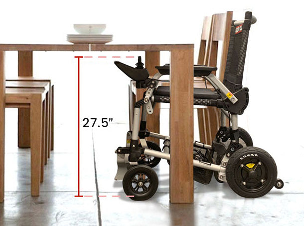 The Journey Zoomer Lightweight Folding Power Chair fits under a wooden dining table with a 27.5-inch height, as shown by a red arrow. Plates are stacked on the tabletop, highlighting the chair’s compatibility for dining use.