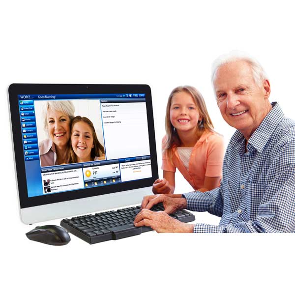 An older man sits at a desk using the WOW! computer by telikin, smiling at the camera. A young girl stands beside him as the simple interface shows a video call with two smiling women.